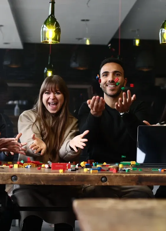 Rosaria Di Donna and Mousa Abdelmaksoud laughing at a wooden table full of Lego bricks, tossing colorful bricks into the air.